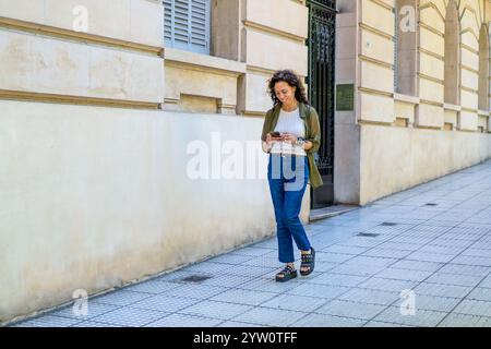 Donna informale che cammina e invia SMS lungo il marciapiede della città Foto Stock