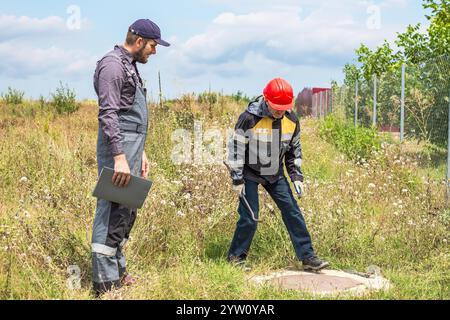 Un appaltatore e un lavoratore in tute con palanchino e chiave regolabile in un'area rurale aprono un pozzo per riparare una rete idrica. Lavoratori municipali. Foto Stock