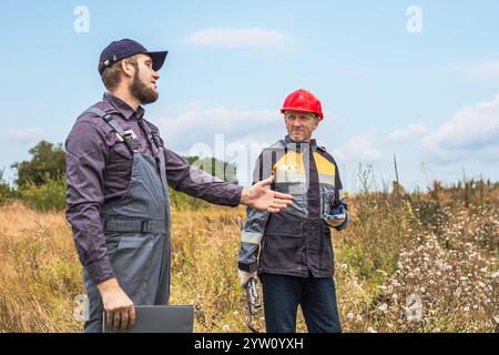 Un appaltatore e un lavoratore in tuta con palanchino e chiave regolabile in un'area rurale discutono del lavoro. Lavoratori municipali, meccanici e idraulici. Foto Stock