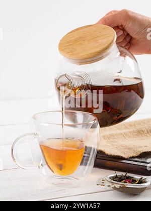 Il tè viene versato da una teiera in una tazza di vetro trasparente. Sfondo bianco in legno. Foto Stock