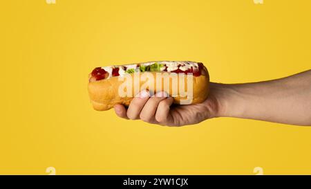 Primo piano di giovane che tiene fresco hot dog con salsa su sfondo arancione, panorama Foto Stock