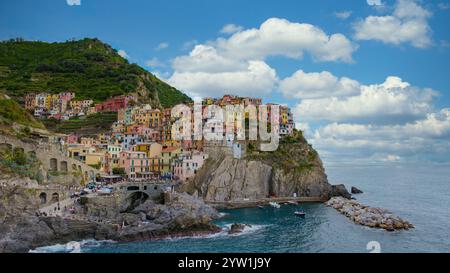 Passeggia per le vivaci strade di Manarola mentre il sole attraversa le nuvole, illuminando le case colorate arroccate sulle scogliere che si affacciano sul tr Foto Stock