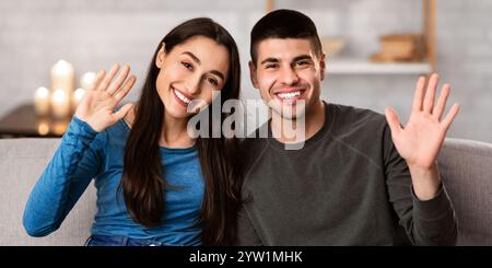 Coppia seduta sul divano a casa e ondeggiante alla macchina fotografica Foto Stock