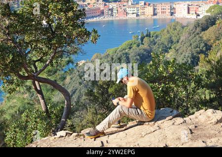 Sestri Levante, Italia - 10 novembre 2024. Escursioni in montagna. Uomo che riposa sulla roccia. Vista sul mare e sulla città. Foto Stock