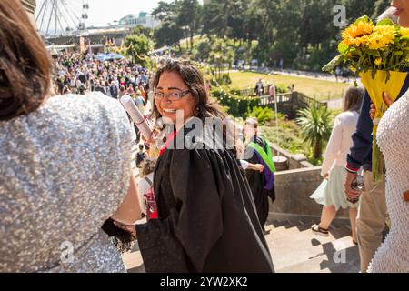 Una donna sorridente in una veste di laurea regge un bouquet di fiori gialli mentre cammina tra una folla di persone all'aperto, Bournemouth, Dorset, Regno Unito Foto Stock