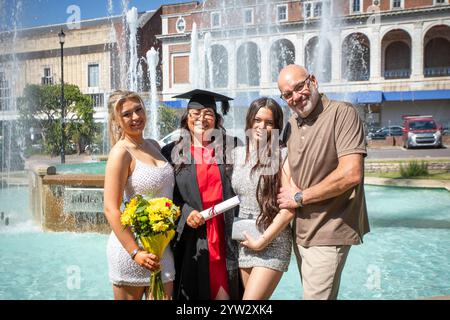 La famiglia celebra il giorno della laurea con una giovane donna sorridente in cappello e abito in possesso di un diploma, accanto a una fontana in una giornata di sole, Bournemouth, Dorset, Regno Unito Foto Stock