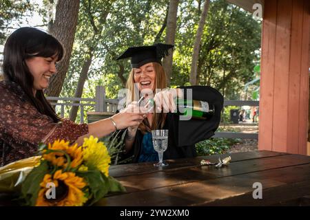 Un laureato sorridente con berretto nero e abito versa champagne in un bicchiere tenuto da un amico a una festa all'aperto, Bournemouth, Dorset UK Foto Stock