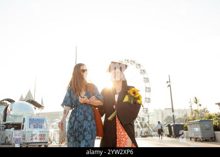 Due donne, una in abito da laurea, che si godono una giornata di sole all'aperto con una ruota panoramica sullo sfondo, Bournemouth, Dorset UK Foto Stock