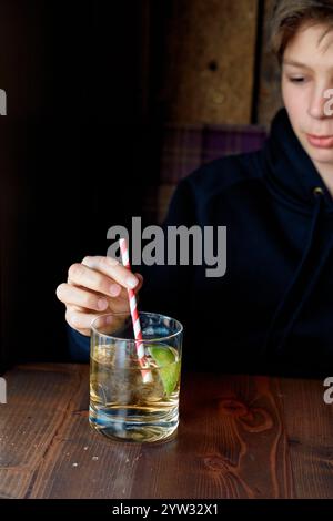 Un adolescente con una felpa scura sorseggia un drink attraverso una cannuccia da un bicchiere trasparente su un tavolo di legno, Edimburgo, Scozia Foto Stock