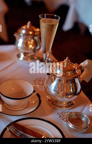 Un elegante ambiente da tavolo con teiera argentata, tazza e piattino, flute di champagne con bibita frizzante e raffinati utensili da pranzo su una tovaglia bianca incontaminata, Edimburgo, Scozia Foto Stock