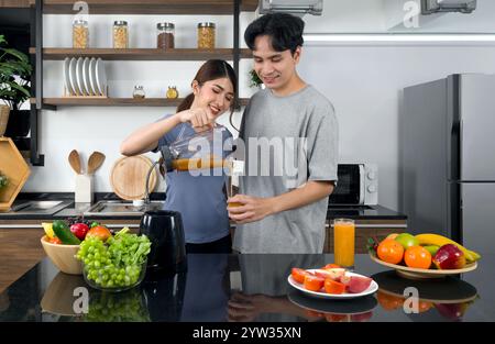 Una giovane donna con un abbigliamento casual versa frullato misto di frutta e verdure dalla macchina per frullare nel bicchiere del suo ragazzo. Gli amanti asiatici trascorrono la mattinata a toge Foto Stock