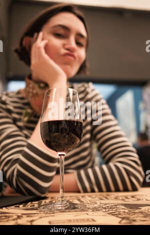 Donna seduta a un tavolo con un bicchiere di vino rosso, che sembra penosa e tentata di bere il vino Foto Stock