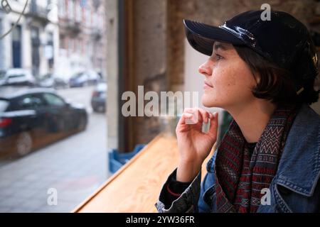 Una giovane donna contemplativa che indossa un berretto nero e una giacca in denim guarda fuori dalla finestra in un bar, perso nel pensiero. Foto Stock