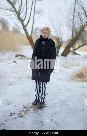 Una giovane donna si trova in un paesaggio innevato, indossando un cappotto nero, pantaloni a righe e stivali, con sciarpa e fascia. Foto Stock