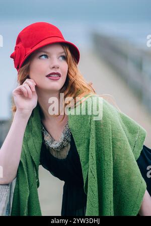 Donna elegante con cappello rosso e scialle verde che guarda attentamente in lontananza. Foto Stock