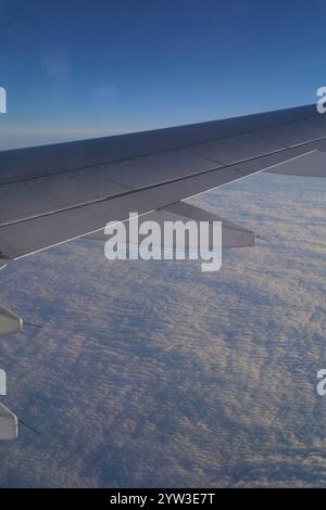 Ala di un aeroplano che vola sopra un paesaggio nuvoloso ad alta quota con cielo azzurro cristallino sullo sfondo. Foto Stock