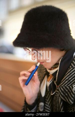 Giovane donna con un cappello nero peloso e un cappotto che svapano pensivamente mentre guarda in basso. Foto Stock