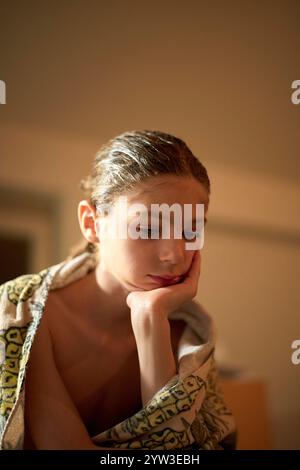 Un giovane contemplativo con i capelli bagnati avvolti in un asciugamano appoggia il mento sulla sua mano Foto Stock