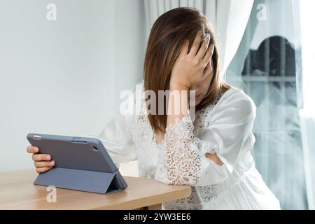 Una donna asiatica in un abito da notte in raso bianco prova dolore agli occhi e fatica mentre legge il suo programma di lavoro su un tablet, facendola stare al passo con lei Foto Stock