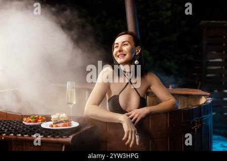 la donna gode di un momento tranquillo in una vasca idromassaggio, circondata dal vapore, assaporando gustosi spuntini e sorseggiando una bevanda. l'atmosfera tranquilla crea una serata perfetta Foto Stock