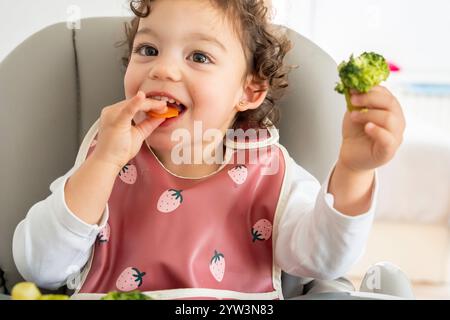 bambina seduta sul suo seggiolone gustando verdure, tenendo in mano broccoli e carote. mangiare sano nell'infanzia, nella cura e nel bene... Foto Stock