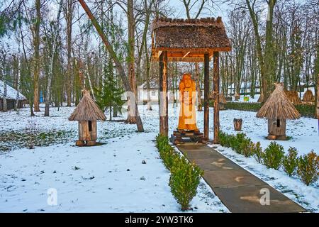 La piccola cappella all'aperto con la scultura in legno intagliato e le antiche case di legno nel parco innevato, Pereiaslav Scansen, Ucraina Foto Stock