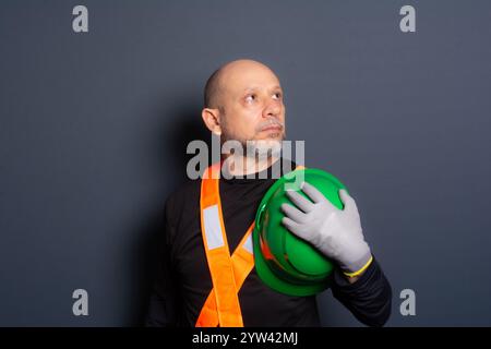 Foto di un impiegato civile con guanti e giubbotto riflettente che tiene il casco verde contro il petto. Isolato su sfondo grigio. Operaio edile. Foto Stock