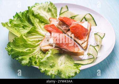 Panini con formaggio fresco e fette di trota salata su un piatto bianco con cetriolo ed erbe aromatiche. Foto di alta qualità Foto Stock