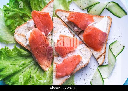 Panini con formaggio fresco e fette di trota salata su un piatto bianco con cetriolo ed erbe aromatiche. Foto di alta qualità Foto Stock