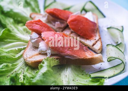 Panini con formaggio fresco e fette di trota salata su un piatto bianco con cetriolo ed erbe aromatiche. Foto di alta qualità Foto Stock