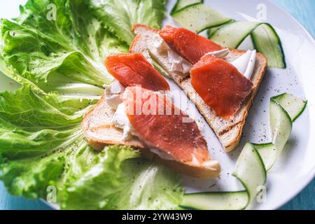 Panini con formaggio fresco e fette di trota salata su un piatto bianco con cetriolo ed erbe aromatiche. Foto di alta qualità Foto Stock