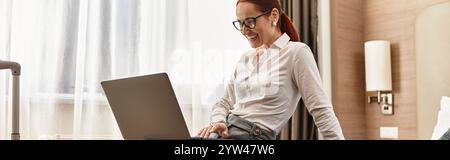 Una donna sorridente si impegna a lavorare dal proprio computer portatile mentre si siede comodamente in una camera d'albergo. Foto Stock