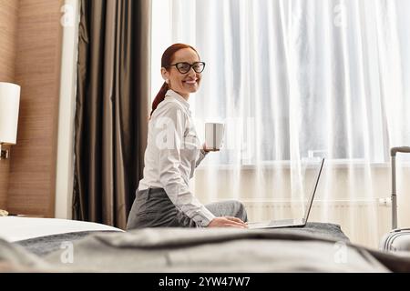 Una giovane donna allegra beve un caffè e lavora con il suo laptop in una camera luminosa e moderna. Foto Stock