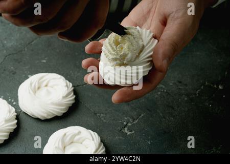Mani che imbettano la panna montata sui nidi di meringa. La superficie a trama scura crea un contrasto rustico con la meringa bianca. Il processo di decorazione di merin Foto Stock