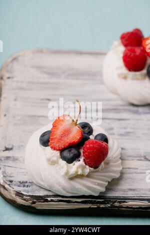 Primo piano di dessert pavlova conditi con fragole fresche, mirtilli e lamponi, disposti su una tavola rustica di legno contro un leggero girone di tè Foto Stock