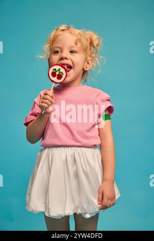 Bambino felice, ragazza con intonaco verde sul braccio dopo la vaccinazione, leccare lecca lecca-lecca su sfondo blu studio. Bambino coraggioso. Foto Stock