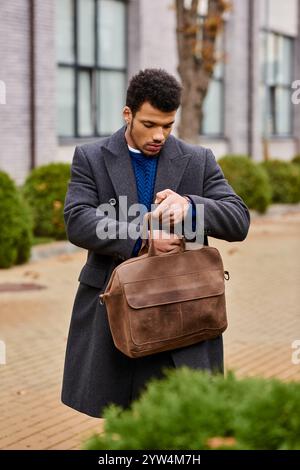 Un giovane alla moda recupera oggetti dalla sua borsa marrone mentre cammina per una strada della città. Foto Stock