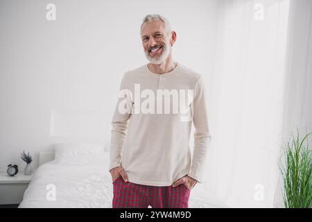 Uomo anziano rilassato in pigiama a quadri che si gode una mattinata luminosa nella sua accogliente camera da letto Foto Stock