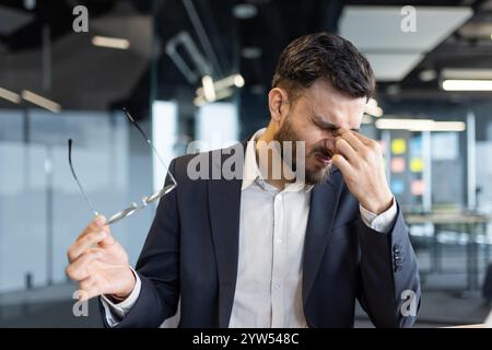 Un uomo d'affari stanco con un vestito si strofina gli occhi tenendo gli occhiali in un ufficio. Sembra stressato o affaticato, indicando un ambiente di lavoro impegnativo. Foto Stock