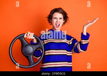 Foto di una giovane ragazza che indossa un maglione blu a righe che tiene il volante le spalle del tappeto arrabbiata dopo un incidente isolato su sfondo arancione Foto Stock