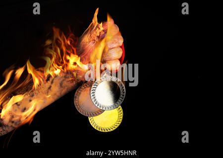 Uomo con medaglie d'oro, d'argento e di bronzo nella mano fiammeggiante su sfondo nero, primo piano Foto Stock