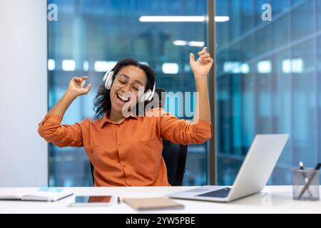 Un allegro lavoratore d'ufficio seduto a una scrivania con un computer portatile, indossando le cuffie e ascoltando musica. La scena trasmette un senso di relax e gioia in un ambiente professionale. Foto Stock