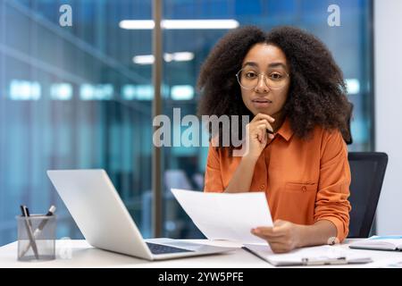 Una donna d'affari concentrata siede a una scrivania in un ufficio moderno, a rivedere i documenti. Il suo laptop è aperto e appare profondamente nel pensiero, incarnando professionalità e concentrazione in un ambiente di lavoro Foto Stock