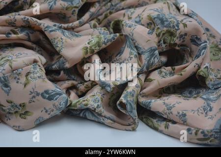 Tessuto estivo insolito e lucido. Il sottile tessuto di seta sintetico beige con fiori blu e foglie verdi si trova su uno sfondo di plastica bianca. Foto Stock