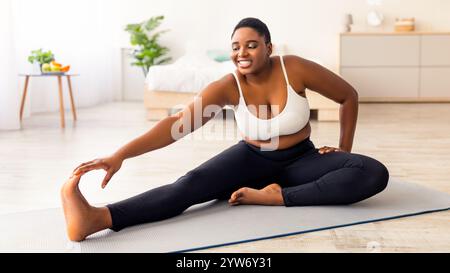 Donna nera curvy che allunga le gambe, si esercita sul tappeto di yoga, leader di stile di vita attivo, cercando di perdere peso a casa Foto Stock