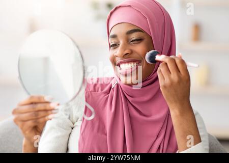 Bella donna musulmana nera in Hijab che guarda in Magnifying Mirror e applica Blush Foto Stock