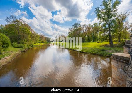 Splendido paesaggio visto da un piccolo ponte sul lungo fiume in un parco pieno di radure verdi, alberi e cespugli al mattino nuvoloso e soleggiato Foto Stock