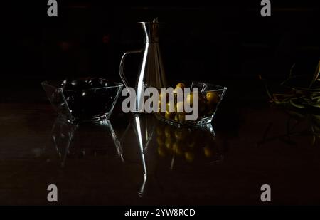 Allestimento scuro e soffice con olive nere e verdi in ciotole di vetro accanto a un barattolo di olio d'argento. Foto Stock