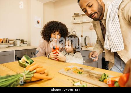 Una famiglia allegra trascorre del tempo di qualità cucinando un pasto insieme nella loro calda cucina. Il bambino sta utilizzando uno smartphone per cercare una ricevuta. Foto Stock