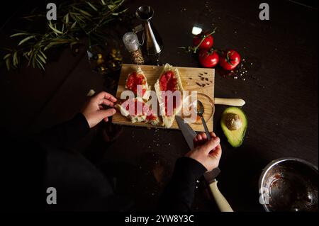 Mani che preparano la bruschetta con pomodoro fresco e avocado su una tavola di legno in un accogliente ambiente di cucina, circondato da ingredienti freschi. Foto Stock